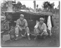 Black and white photo of two young Navajo Code Talkers sitting near their camp outside.  Both face the camera.