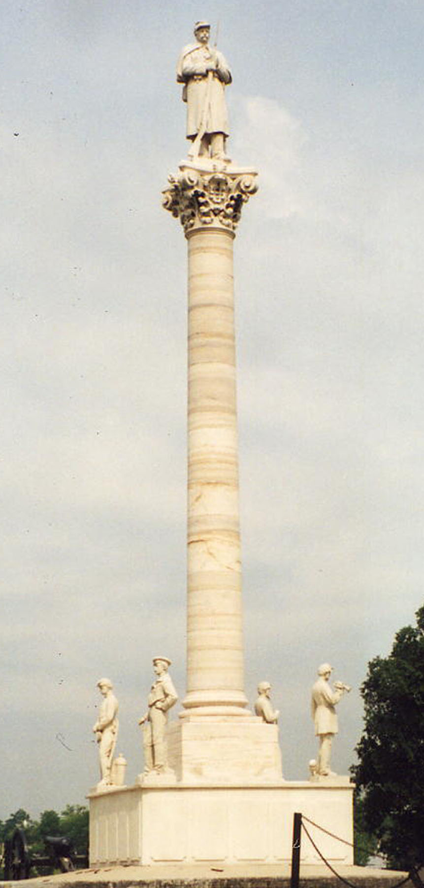 Photo of a tall white marble monument with four small male figures at each corner of the square base. A larger male figure stands at the top of the pillar.