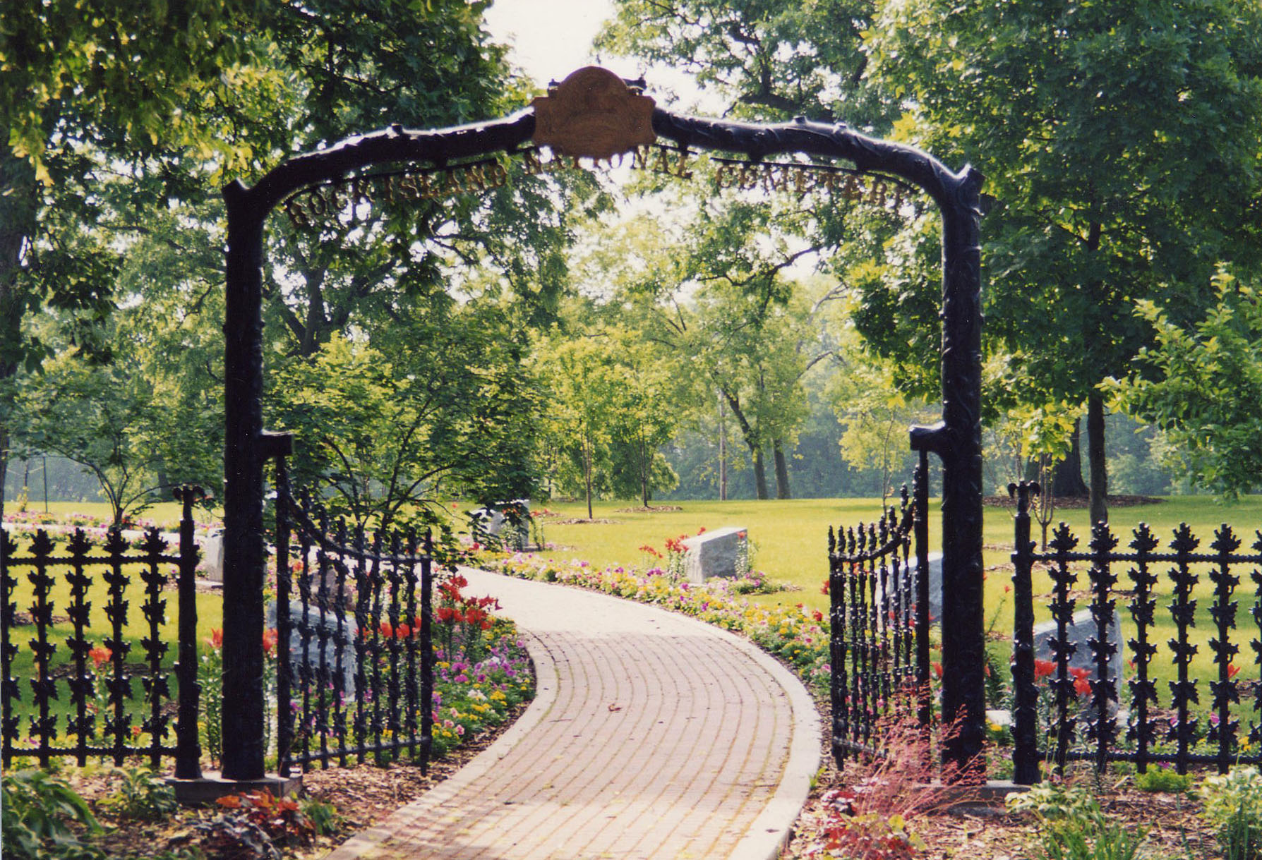 Photo of a tall black iron arch connect to a mid-size fence.  An ivory brick walkway leads into the cemetery through an opened entrance gates.