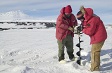 Two people in red parkas drilling through snow and ice.