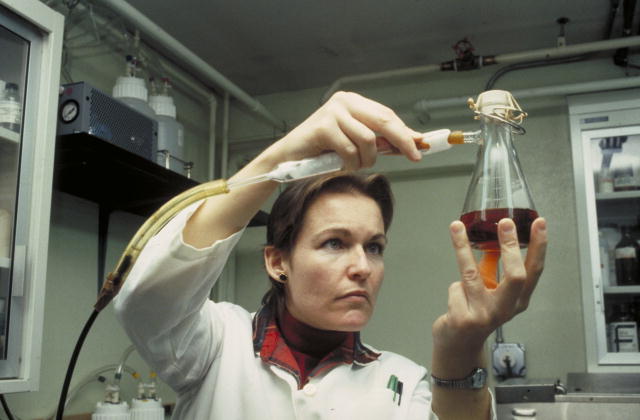 A female USDA scientist is holding a beaker of liquid in one hand while holding a tube attached to a hood over the beaker opening. She is analyzing this sample for contamination.