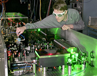 Scientist wearing dark goggles turns knob to adjust a green laser reflecting off mirrors on a laboratory table.
