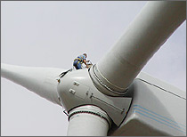 Photo of a man who appears tiny atop the hub of a massive wind turbine.
