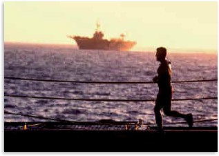 A Marine jogs on the flight deck of an amphibious transport dock with an amphibious assault ship in the background.