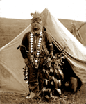 This 1915 photo shows a young Native American dancer in traditional Lakota dance attire complete with dance bells and feather headdress.  Note the young man's mirror stole, which originated from the days of the fur trade.  It's unusual that this man lives in a U.S. military tent called a Sibley.  When buffalo hides became scarce, the U.S. Government supplied canvas tents and canvas material to make shelters.  These tents were very warm, even in the dead of winter, if a small fire was kept constantly burning.  -- select to view larger image.