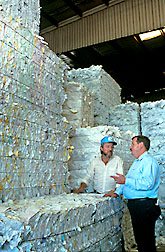 Photo of two men standing in front of stacks of paper for recycling.